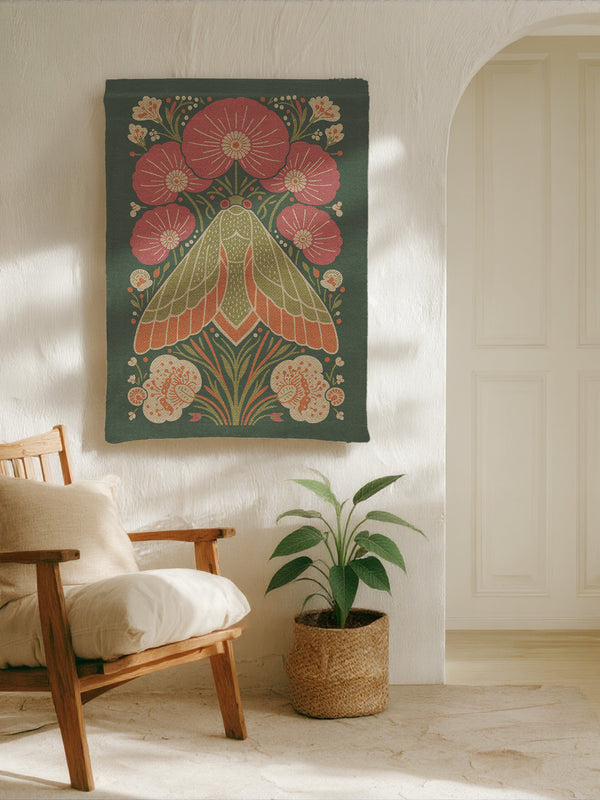 A rectangular woven wall tapestry with a rich, dark green background. The central design is a large, stylized moth with intricate wings in shades of orange, cream, and muted green. The moth is surrounded by a symmetrical arrangement of folk-art style flowers and botanicals in vibrant pink, coral, and cream. The entire piece has a visible, high-quality woven texture. This polyester and cotton blend textile art measures 26x36 inches and includes a rod pocket on the black backing for easy hanging.
